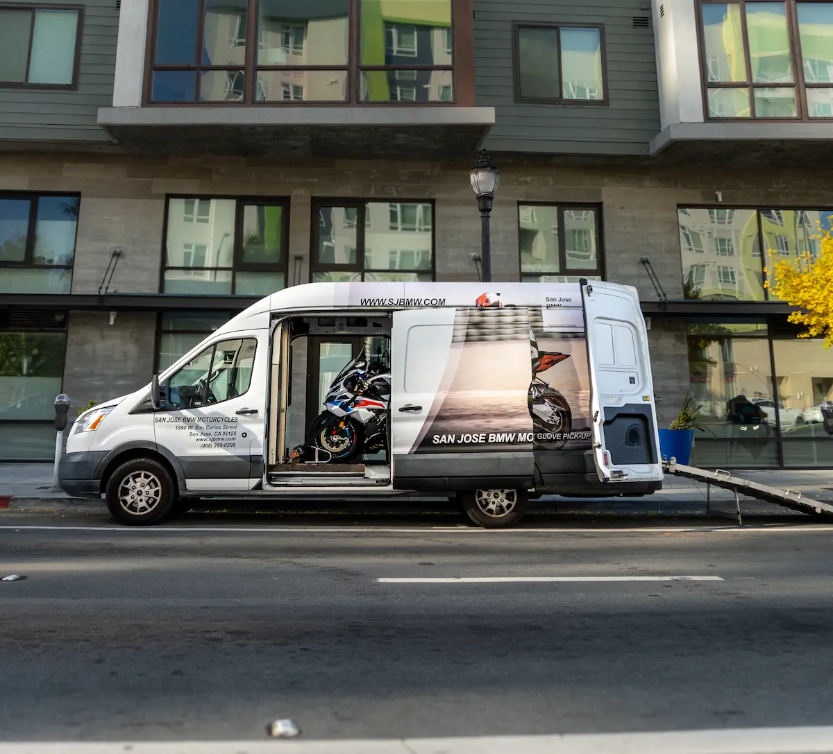 San Jose BMW Motorcycles branded Ford Transit white cargo van parked on a city street, hauling a BMW sport motorcycle inside with its side sliding door open.